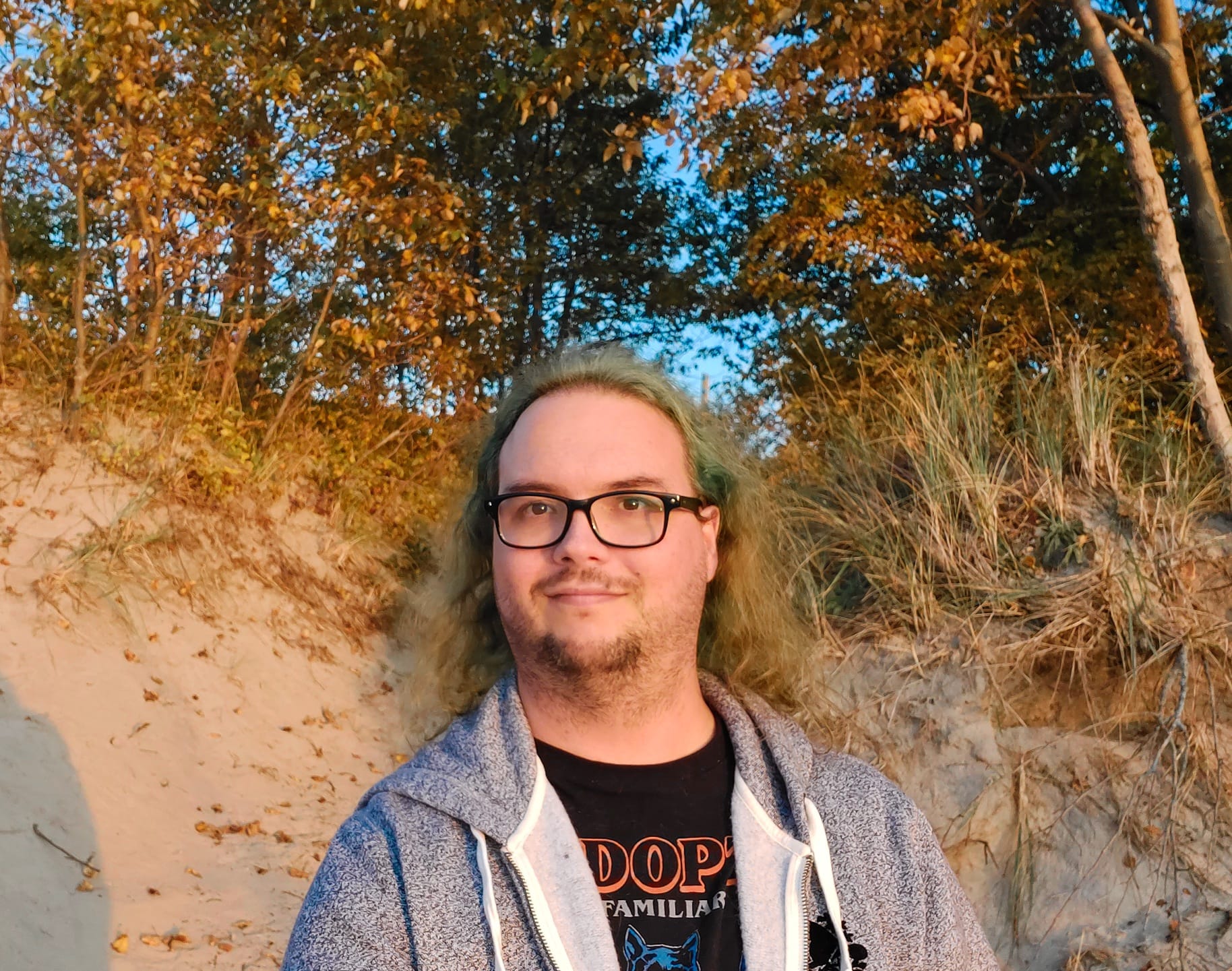 A man with green hair wearing a swetter stands in front of a sand dune with trees on it, the sun shines on him and half the dune